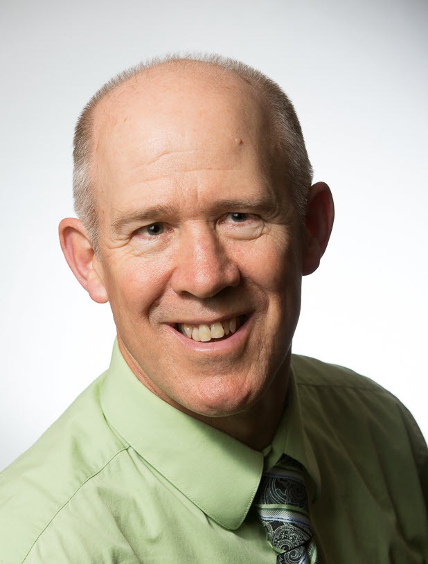 Smiling man in a light green shirt and patterned tie against a plain background, conveying a professional and approachable demeanor.