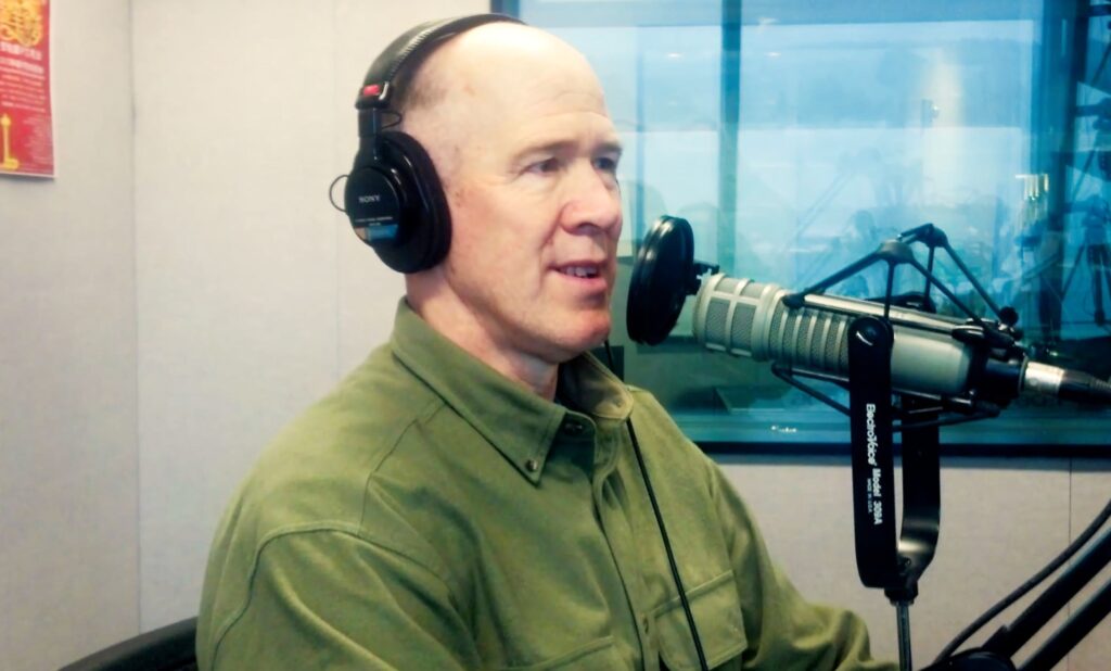 Man wearing headphones speaks into a microphone in a radio studio, conveying a conversation or interview context.